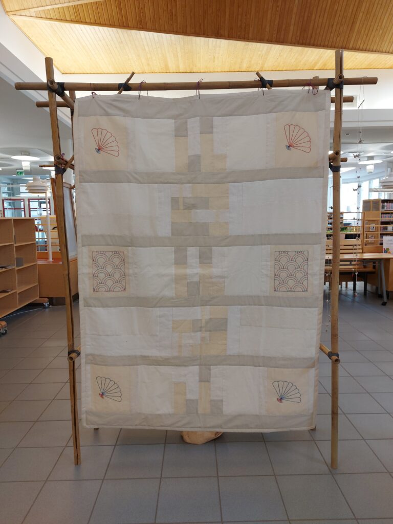 Telineeseen ripustettu peitto, jossa on kirjomalla koristeltuja tilkkuja.