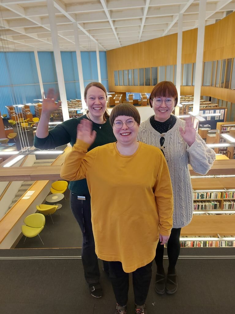 Kolme ihmistä seisoo kirjaston parvella ja vilkuttaa